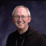 A smiling older man with glasses, short gray hair, and wearing a black robe with a chain necklace, posed in front of a dark blue background.